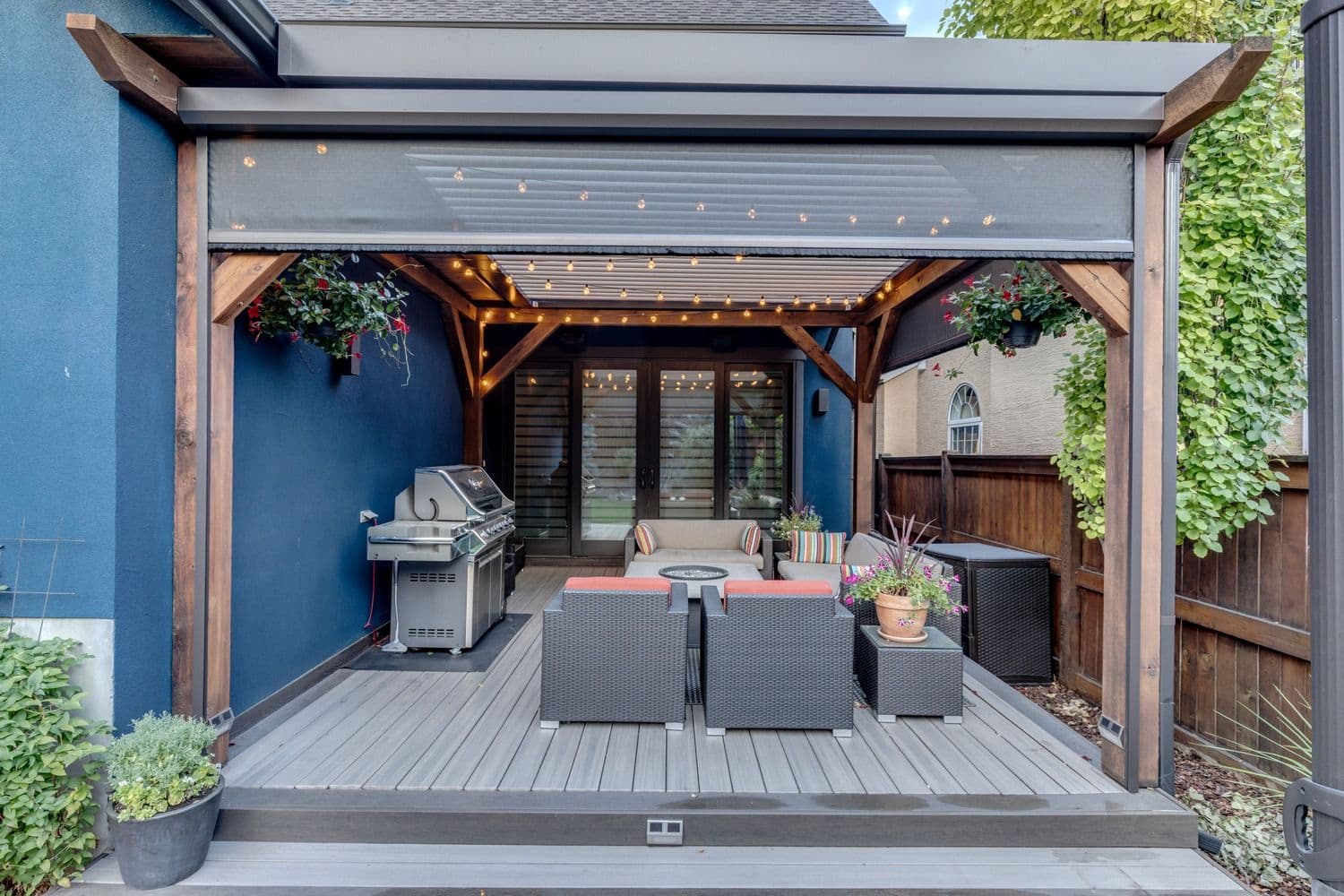 Modern backyard patio with retractable louvered pergola, gas grill, lounge seating with orange cushions, fire pit table, string lights, and potted flowers surrounded by blue walls and ivy-covered fencing